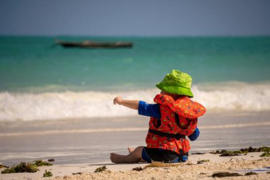 Küçük beyaz çocuk, okyanusta bir yaz plajında, deniz dalgalarının arka planında can yeleği ve parmağıyla işaret eder.