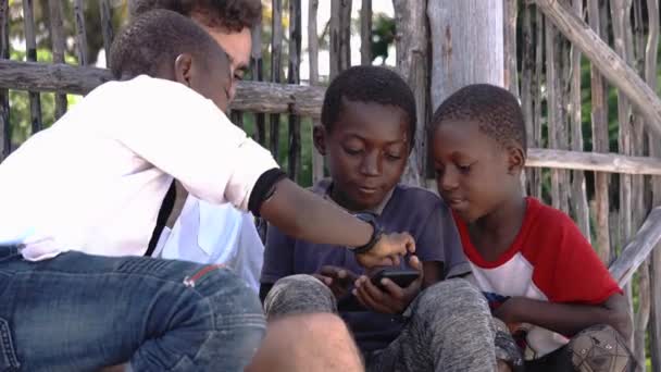 Tanzania, Zanzibar - januari 2020: Ung kaukasisk man som leker med svarta afrikanska pojkar som visar spel på sin smartphone — Stockvideo