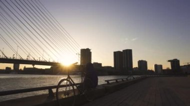 Bisikletli bir adam Erasmus Köprüsü yakınlarındaki Embarkment 'ta oturuyor ve Rotterdam, Hollanda' da Sunset saatinde marihuana ile esrar içiyor.