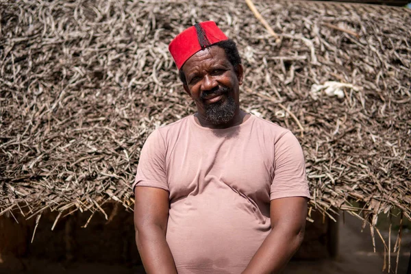 Um homem mais velho africano em vermelho muçulmano Taqiyyah Fez chapéu posando com um pau para as pessoas coxos no quintal Perto da cabana básica com telhado coberto em pequena aldeia remota na Tanzânia, ilha de Pemba, Zanzibar — Fotografia de Stock