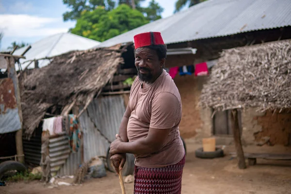 Um homem mais velho africano em vermelho muçulmano Taqiyyah Fez chapéu posando com um pau para as pessoas coxos no quintal Perto da cabana básica com telhado coberto em pequena aldeia remota na Tanzânia, ilha de Pemba, Zanzibar — Fotografia de Stock