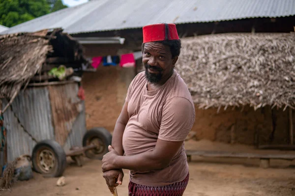 Um homem mais velho africano em vermelho muçulmano Taqiyyah Fez chapéu posando com um pau para as pessoas coxos no quintal Perto da cabana básica com telhado coberto em pequena aldeia remota na Tanzânia, ilha de Pemba, Zanzibar — Fotografia de Stock