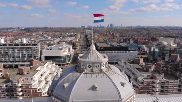 Ave aérea Vuele lejos vista sobre una bandera en la parte superior del Kurhaus de Scheveningen Situado en la costa holandesa. Larga playa de arena un paseo marítimo y un muelle en el Mar del Norte popular destino turístico — Vídeo de stock