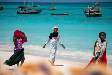 ZANZIBAR, TANZANIA - 2020 HAZİRAN: Üç genç kız, Nungwi, Zanzibar 'daki Okyanusta Turkuaz Su ve Dhow Tekneleri ile sahilde