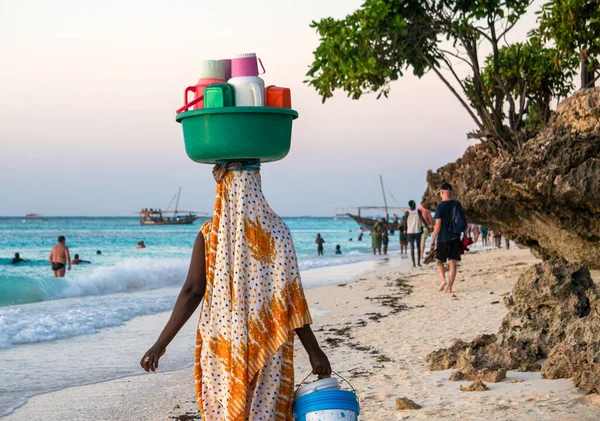 Africké místní ženy nesoucí poháry, plastové konvice a termosky s čajem a kávou na prodej na pláži na její hlavu na pláži s tyrkysovou vodou v Nungwi, Zanzibar, Tanzanie — Stock fotografie