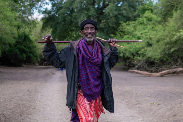 TANZÂNIA, ÁFRICA ORIENTE - JANEIRO 2020: O homem Maasai mais velho com roupas e armas tradicionais está caminhando na savana pela estrada de cascalho cercado de árvores de acácia verdes — Fotografia de Stock