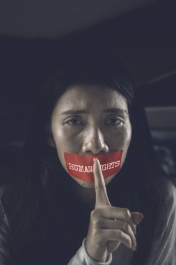 Asian girl gesturing silence while sitting in the dark room with her mouth covered by human rights text on a red tape clipart