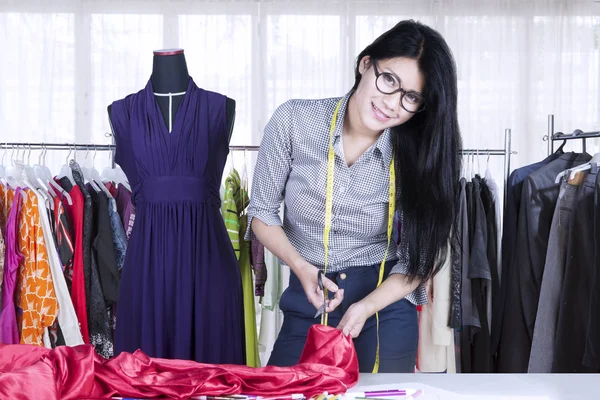Picture of a pretty seamstress cutting a fabric by using scissor while standing in the workplace