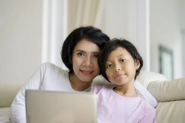 小女孩学习使用笔记本电脑与她的母亲 而看着相机和坐在沙发上 — 图库照片
