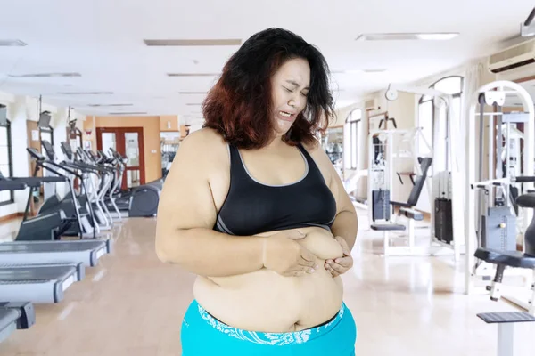 Image of sad obese woman pinching her belly fat while standing in the gym center