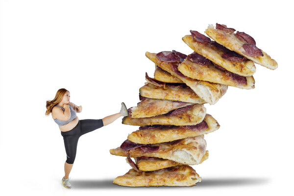 Picture of beautiful fat woman wearing sportswear while kicking a pile of pizzas, isolated on white background