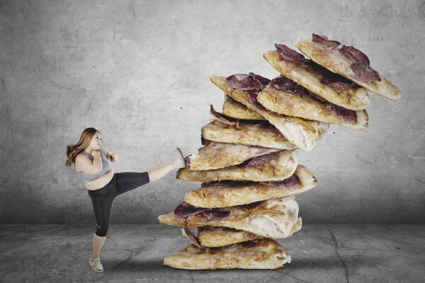 Portrait of obese woman wearing sportswear while kicking a pile of tasty pizzas