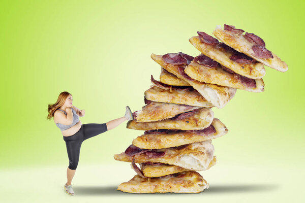 Image of overweight woman wearing sportswear while kicking a pile of pizzas with green screen background