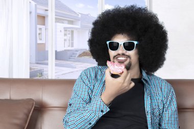 Kıvırcık saçlı genç Afro man kanepede güneş gözlüğü takıyor süre evde donut yemek