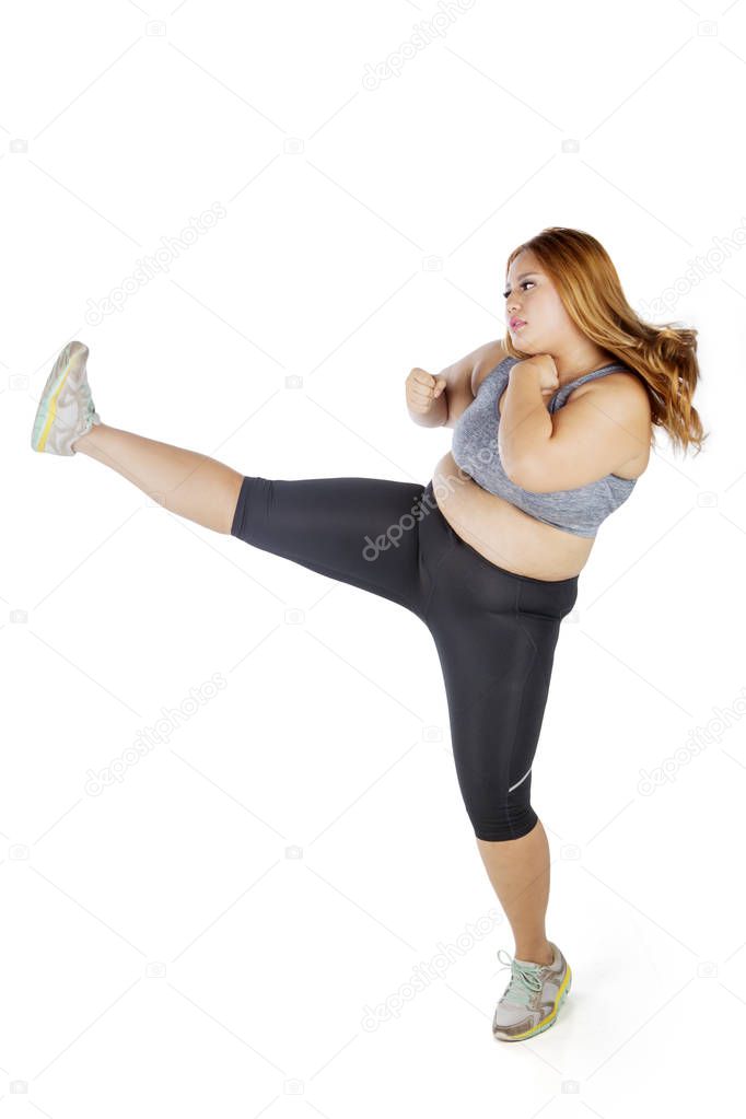 Portrait of pretty overweight woman wearing sportswear while doing a workout by kicking, isolated on white background