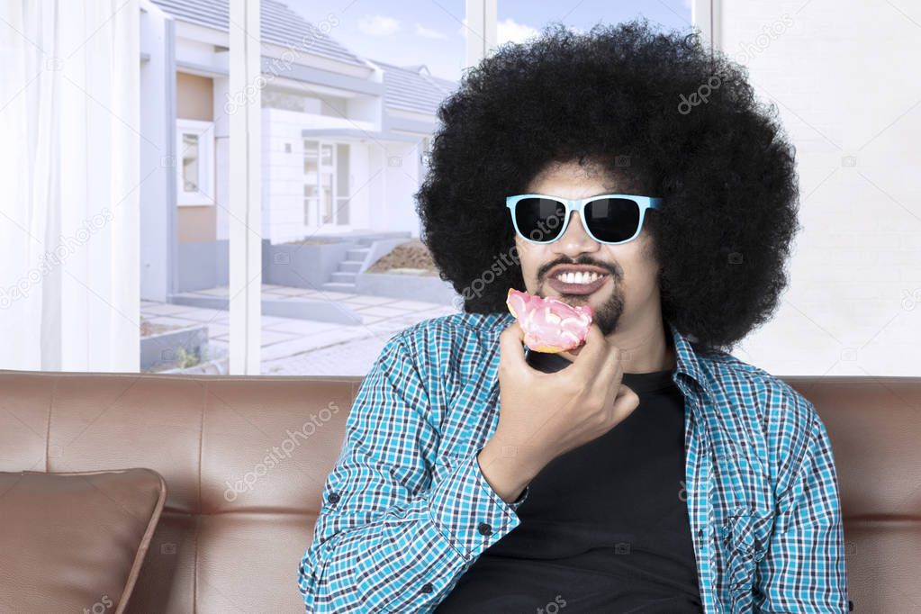 Young Afro man with curly hair, eating donuts at home while wearing sunglasses on the sofa