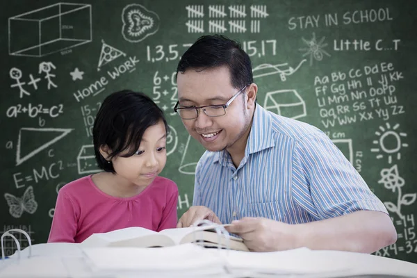 一个小女孩的图片学习阅读一本书与她的老师 而坐在教室里 黑板上的涂鸦 — 图库照片