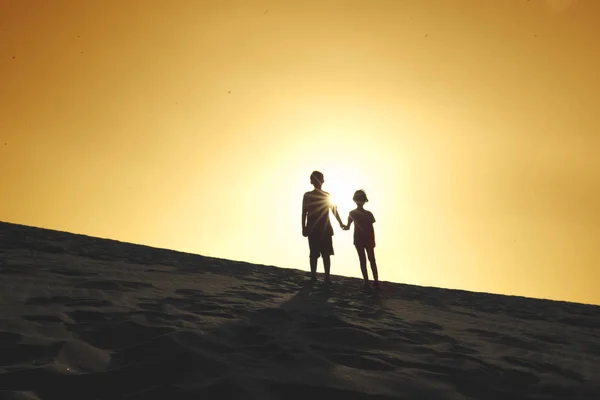 人の子供が手を繋いで一緒に立っている間 砂丘の日没時のシルエット ビーチで撮影します — ストック写真