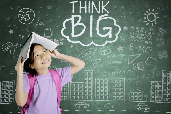 Portrait of pensive schoolgirl looking at word of think big on a chalkboard while putting a book on her head