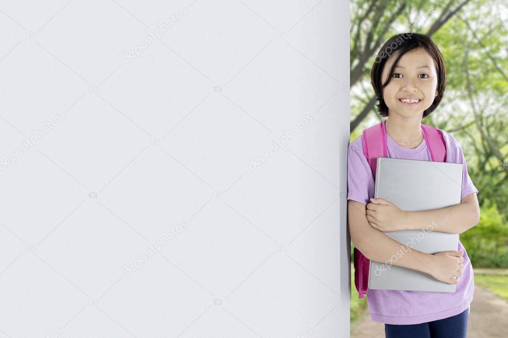 Picture of Asian schoolgirl smiling at the camera while holding a laptop and leaning on the wall