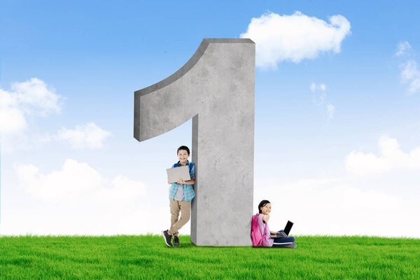 Picture of two elementary students leaning on a rock shaped number one while studying with laptop on the meadow