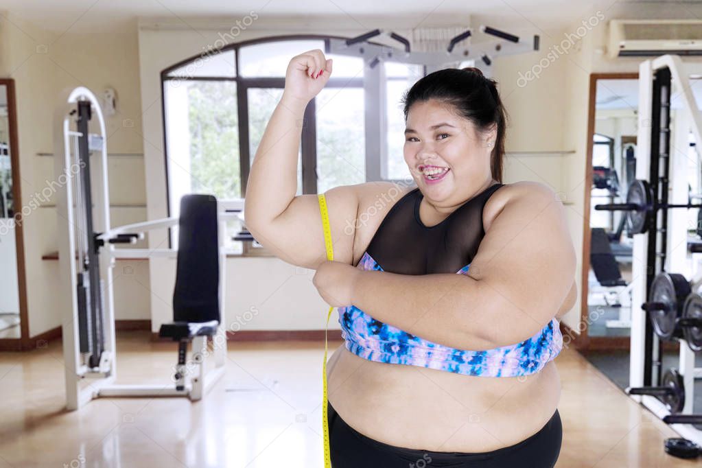 Picture of overweight woman looks happy while measuring her bicep and standing in the gym center