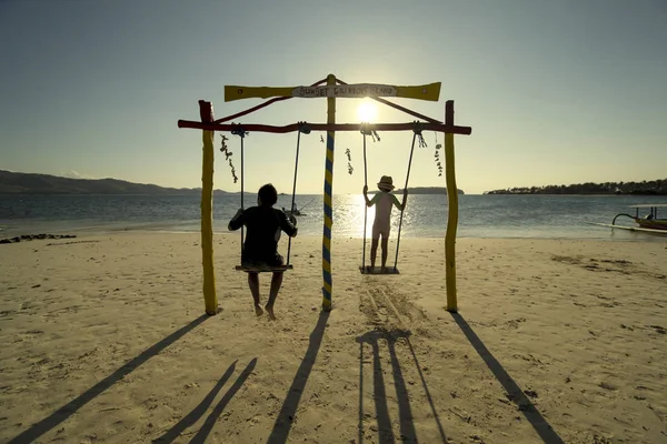 两个孩子在海滩上玩秋千的剪影 在日落时享受海景 在龙目岛拍摄 — 图库照片