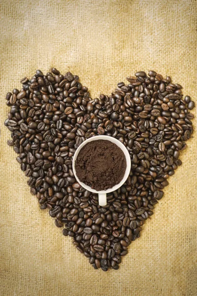 Coffee beans shaped heart symbol with a cup full of ground coffee on the table