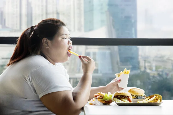 在餐桌上吃汉堡包的胖女人的侧面欣赏法式薯条 — 图库照片