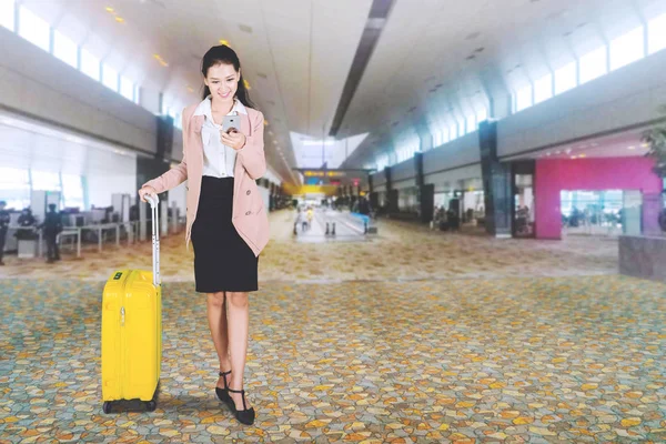 在机场大厅与手提箱站在一起使用智能手机的快乐女商人的图片 — 图库照片