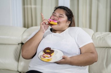 Overweight woman sitting on the couch while eating a plate of donuts. Shot at home clipart