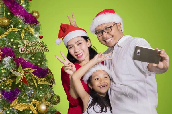 小女孩的图片与她的父母采取自拍照片一起使用智能手机附近的圣诞树 — 图库照片