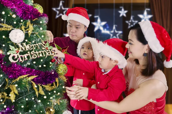 快乐的年轻父母和他们的孩子一起装饰圣诞树 而在家里戴着圣诞帽 — 图库照片