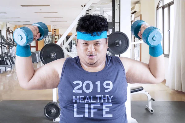 Image of obese man is lifting two barbells in the gym center with text of 2019 healthy life on his shirts
