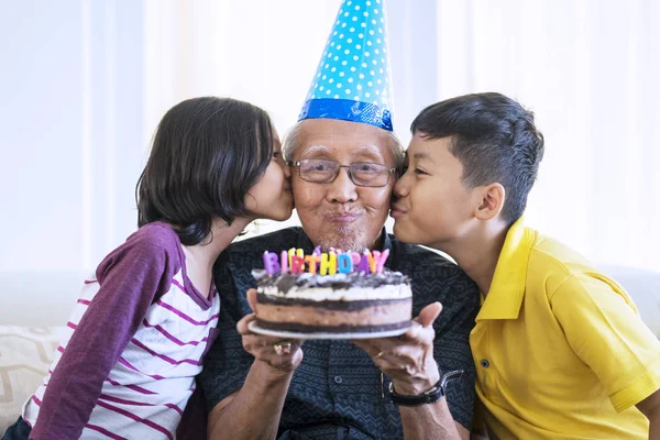 Cheerful old man celebrating his birthday while holding a birthday cake and kissed by grandchildren at home