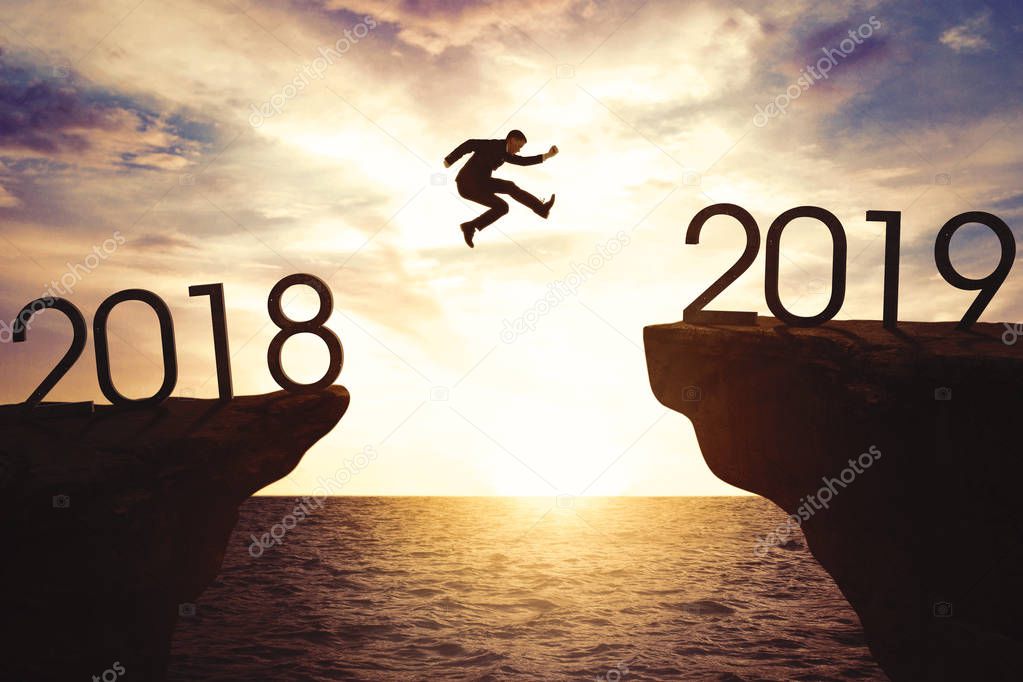 American businessman jumping gap on the cliff with numbers 2018 and 2019. Shot at sunrise time