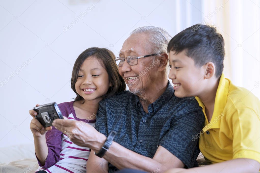 Portrait of happy senior man and his grandchildren holding a mobile phone together on the sofa at home