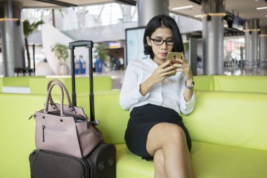 Kadın girişimci bir bavulla Havaalanı dinlenme odasında otururken bir smartphone kullanarak