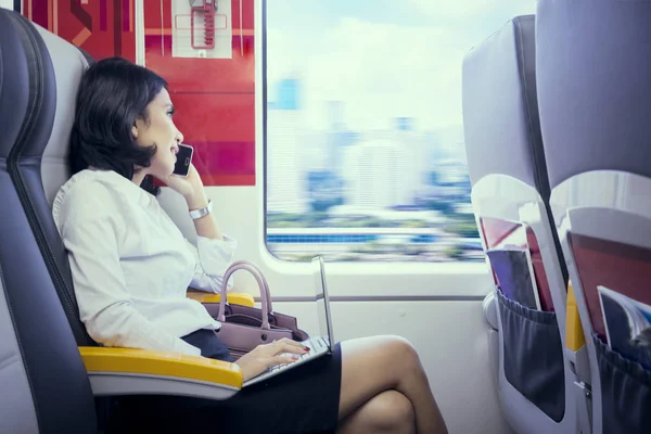 在机场火车上使用笔记本电脑时在手机上讲话的年轻女商人 — 图库照片