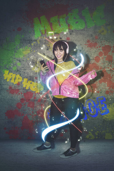 Asian female dancer listening music from her phone while standing with colorful light flow in the studio