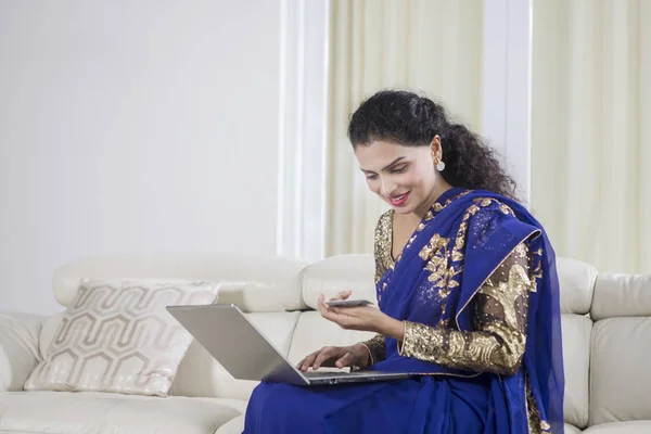 漂亮的印度女人坐在家里的沙发上 一边使用笔记本电脑和信用卡 — 图库照片