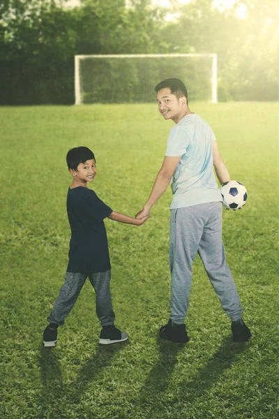 サッカーのフィールドで彼の息子が付いている球を運んでいる間カメラを見て若い父親の後姿 — ストック写真