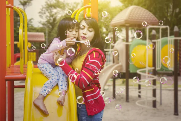 若い女性と彼女の娘が遊び場で一緒にプレーしながらシャボン玉を吹く — ストック写真