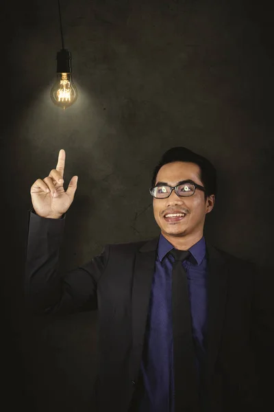 店長の若い男性の肖像画は 暗い部屋で明るい電球を指差しながら幸せそうに見える — ストック写真