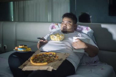 Sağlıksız yaşam tarzı kavramı: Asya obez man yemek ıvır zıvır yiyecek uyku önce yatakta Tv izlerken