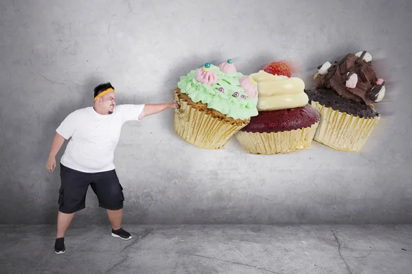 Picture of an obese man refusing to eat sweet food by punching cupcakes in the studio with angry expression