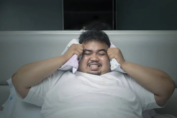 Sleeping disorder concept: Angry obese man unable to sleep during closing his ears with a pillow