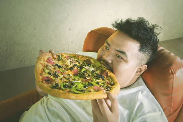 不健康的生活方式概念 年轻的胖子在沙发上放松 同时吃一个大比萨饼与暴饮暴食的表情 — 图库照片