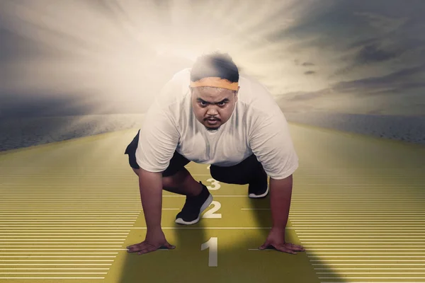 Picture of an Asian fat man ready to run while kneeling above measuring tape. Shot with sunlight background