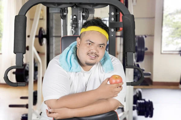 图片是一个年轻的肥胖男子坐在运动机上拿着一个新鲜的苹果 在健身中心被枪杀 — 图库照片